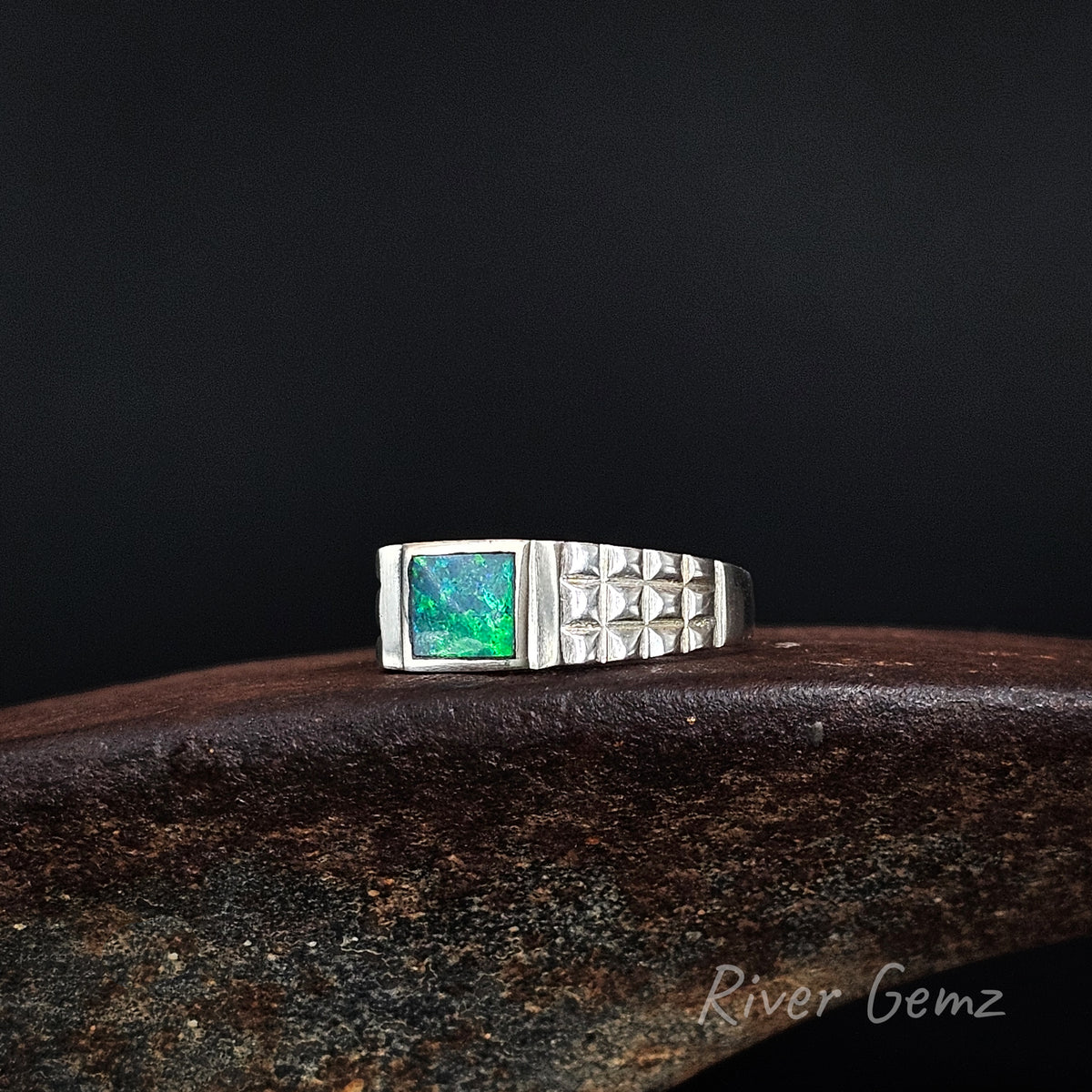 Vibrant green and blue rectangular opal inlaid in men's silver ring with checker plate tapered shoulders on a weathered shoe anvil and black background.