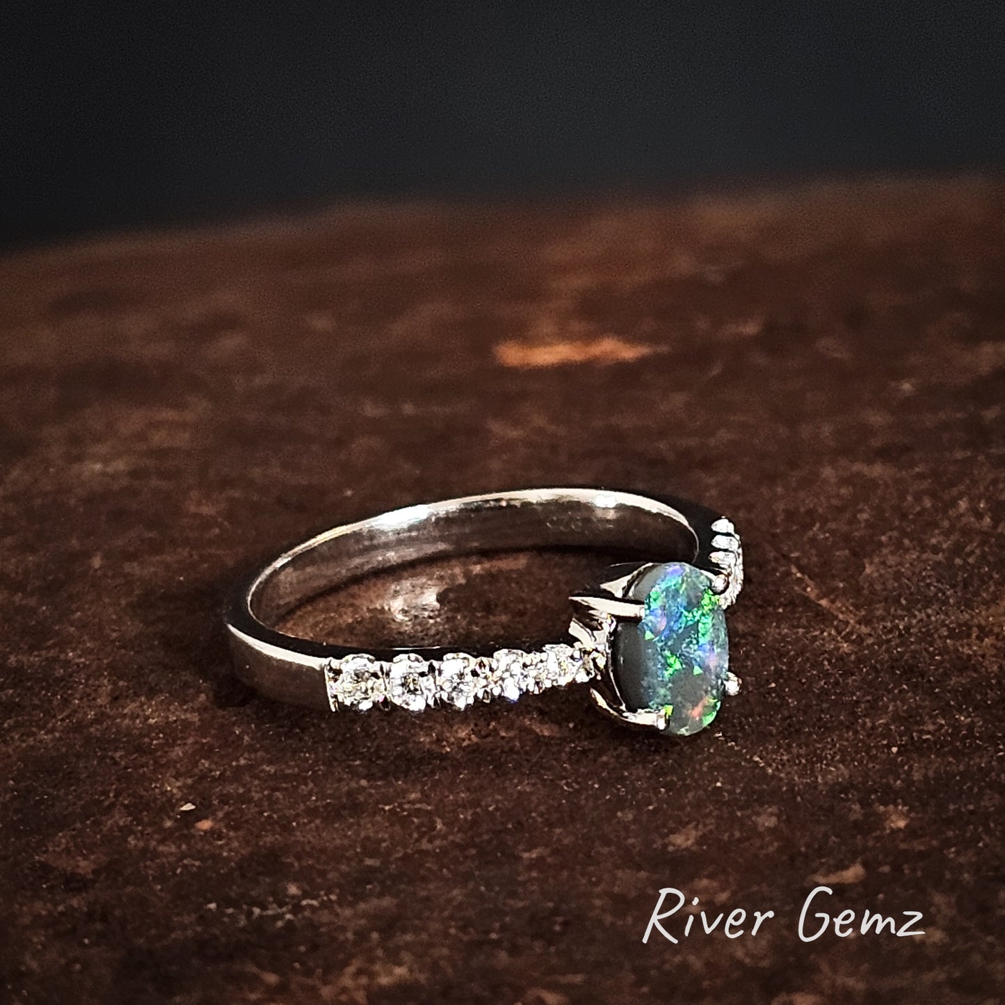 Silver ring with a green gemstone on a dark brown surface, branded 'River Gemz'.