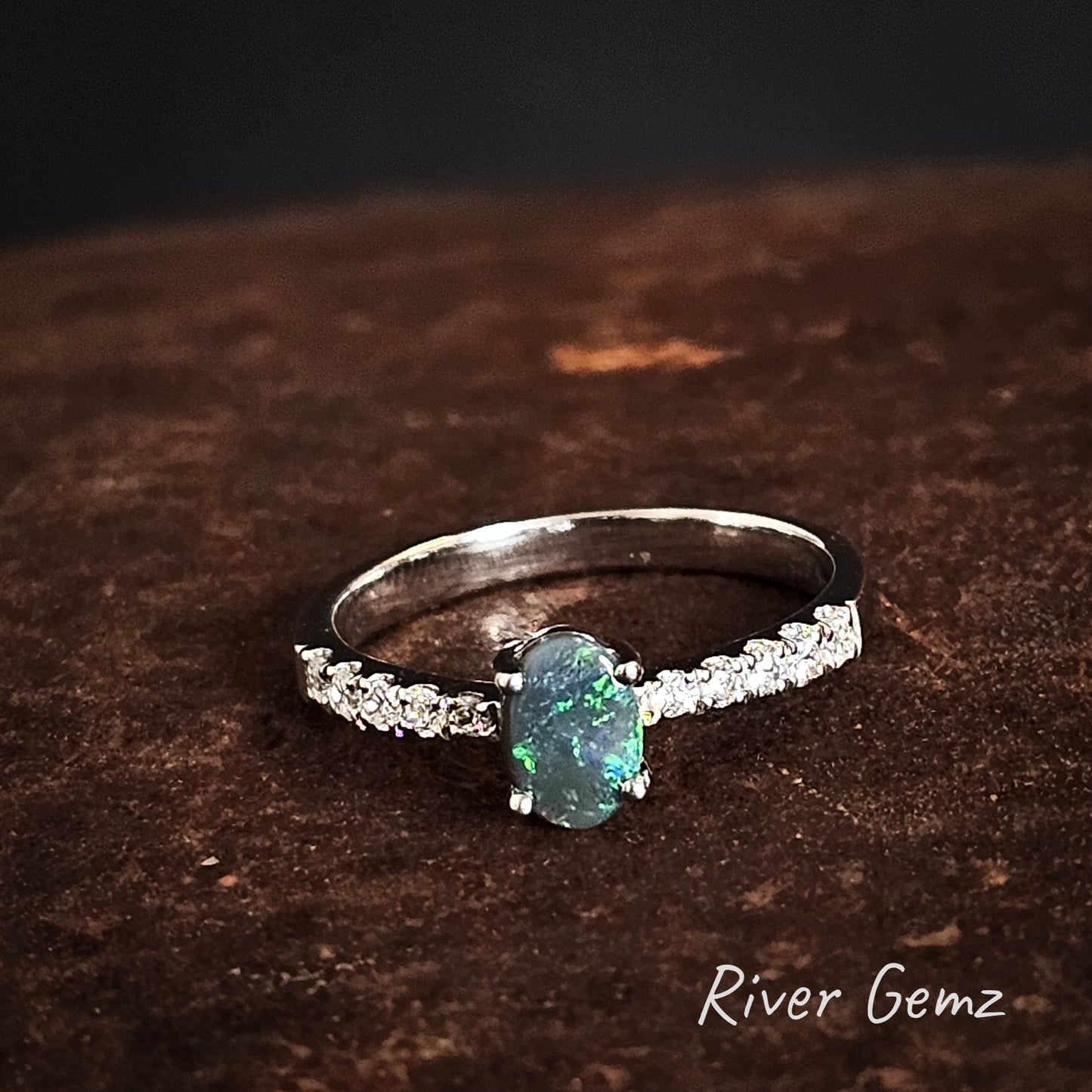 Silver ring with a green opal and 5 white topaz stones on each shoulder  on a dark brown surface, branded 'River Gemz'.