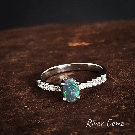 Silver ring with a green opal and 5 white topaz stones on each shoulder  on a dark brown surface, branded 'River Gemz'.