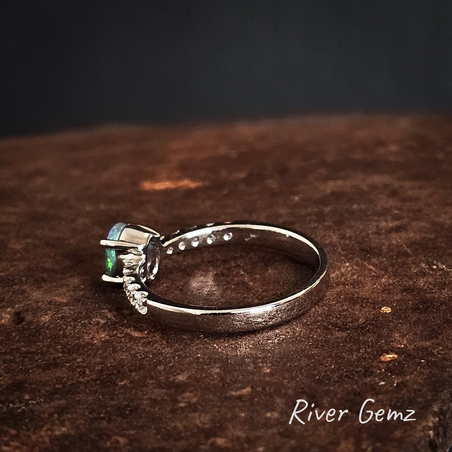 Side view of the silver ring with a green gemstone on a textured brown surface, branded 'River Gemz'.