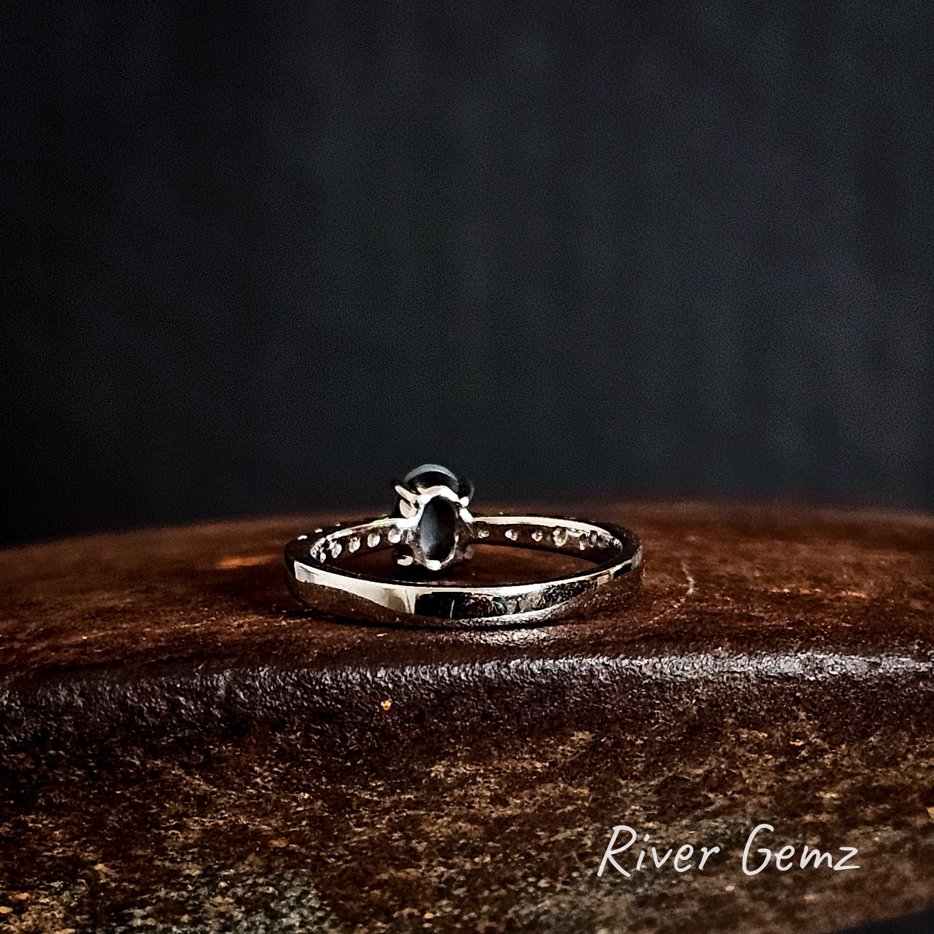 Rear view of the silver ring shows the dark base tone of the black crystal opal on a rust coloured surface, featuring 'River Gemz' branding.