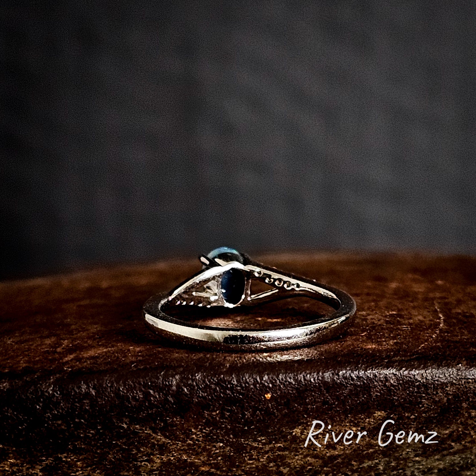 Rear of silver ring shows the dark base tone of the inserted black opal on a dark surface with 'River Gemz' branding.