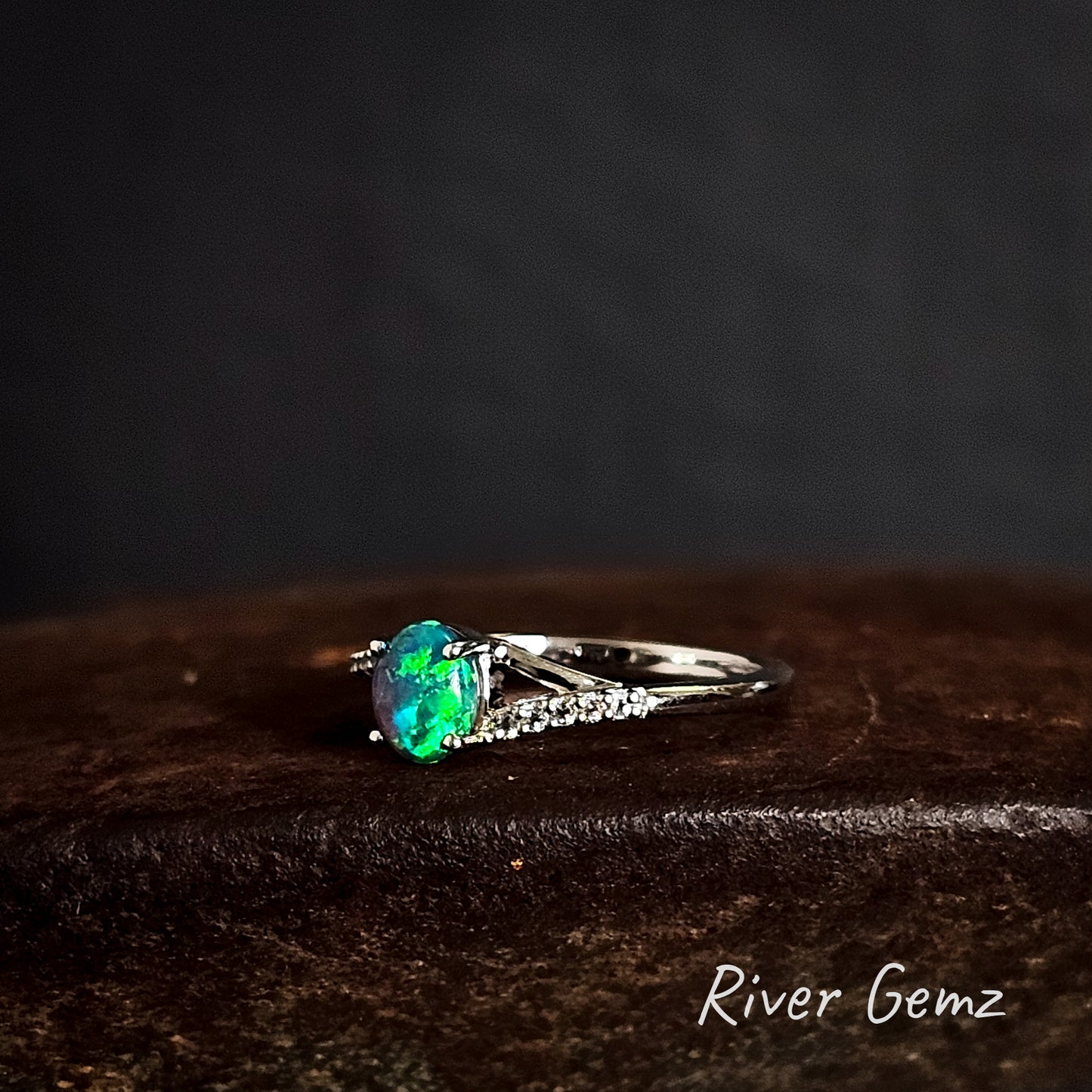Ring with an oval shaped green coloured black opal on a dark brown surface, branded 'River Gemz'.