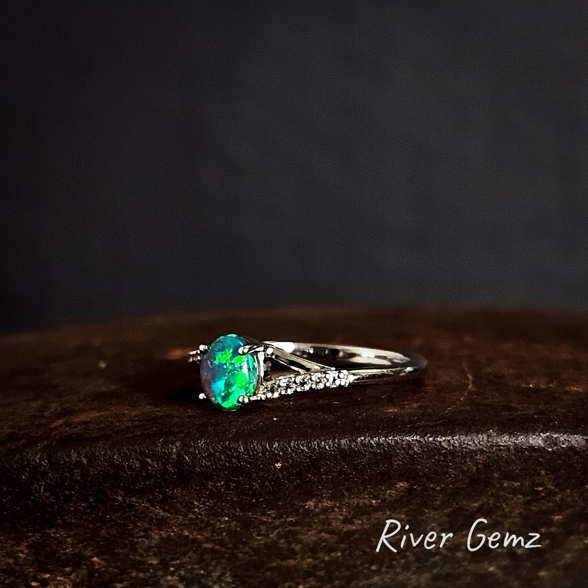 Ring with an oval shaped green coloured black opal on a dark brown surface, branded 'River Gemz'.