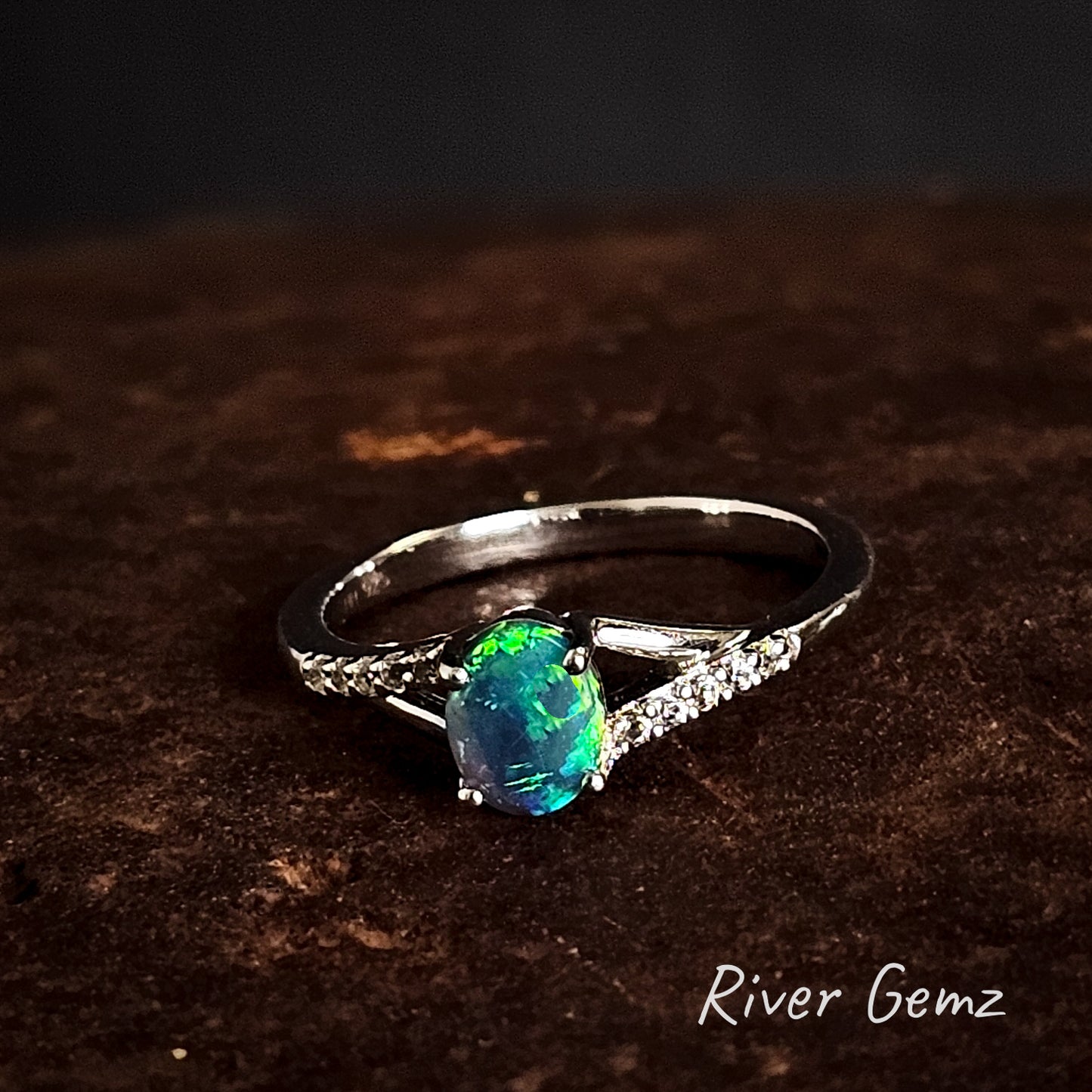 Silver ring with a black crystal opal and white topaz stones on a dark brown surface, branded 'River Gemz'.
