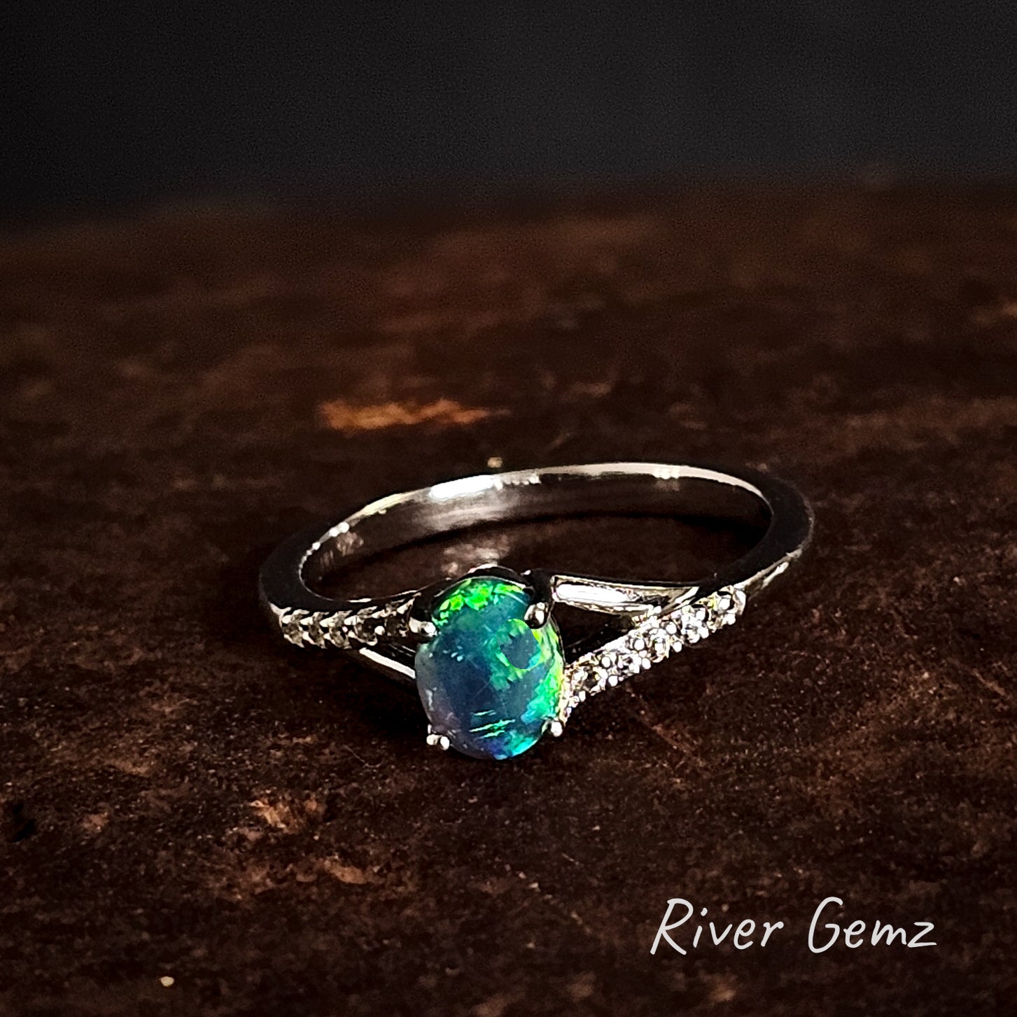 Silver ring with a black crystal opal and white topaz stones on a dark brown surface, branded 'River Gemz'.