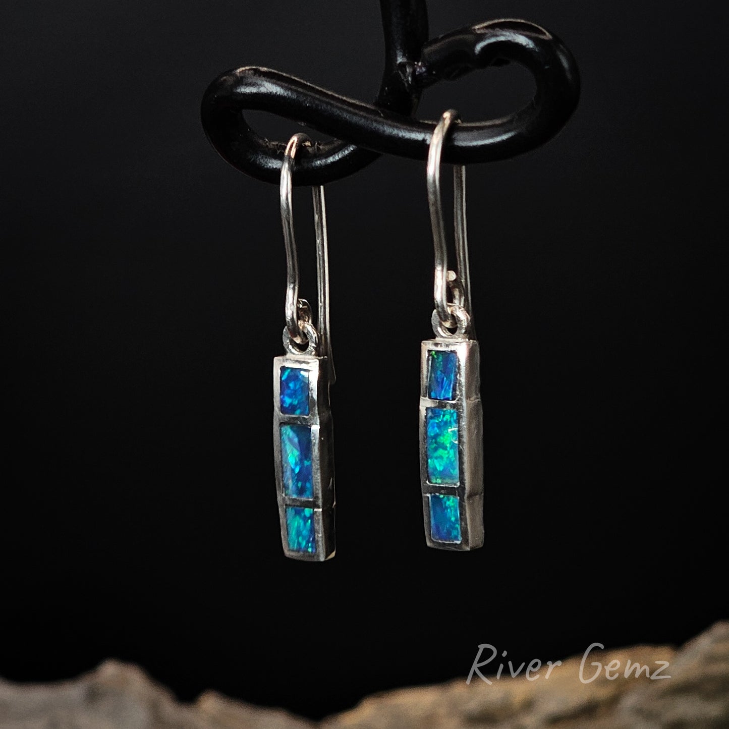 Pair of silver earrings with blue gemstones on a black background, branded 'River Gemz'.