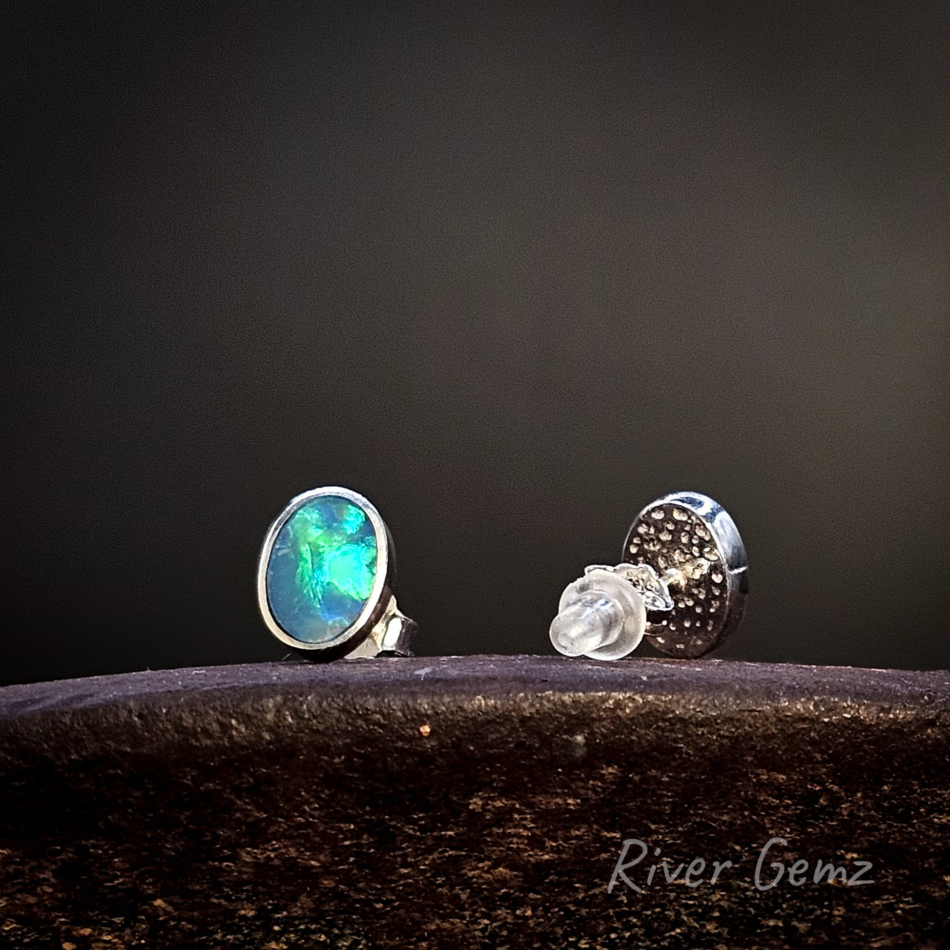 Pair of sterling silver studs with blue and green opals on a dark surface with a black background, featuring 'River Gemz' branding.