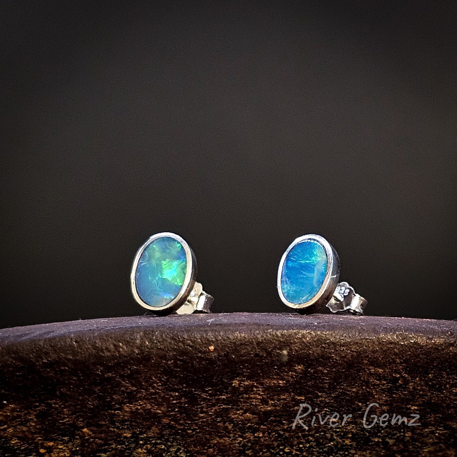 Pair of blue gemstone stud earrings on a dark background with 'River Gemz' branding.