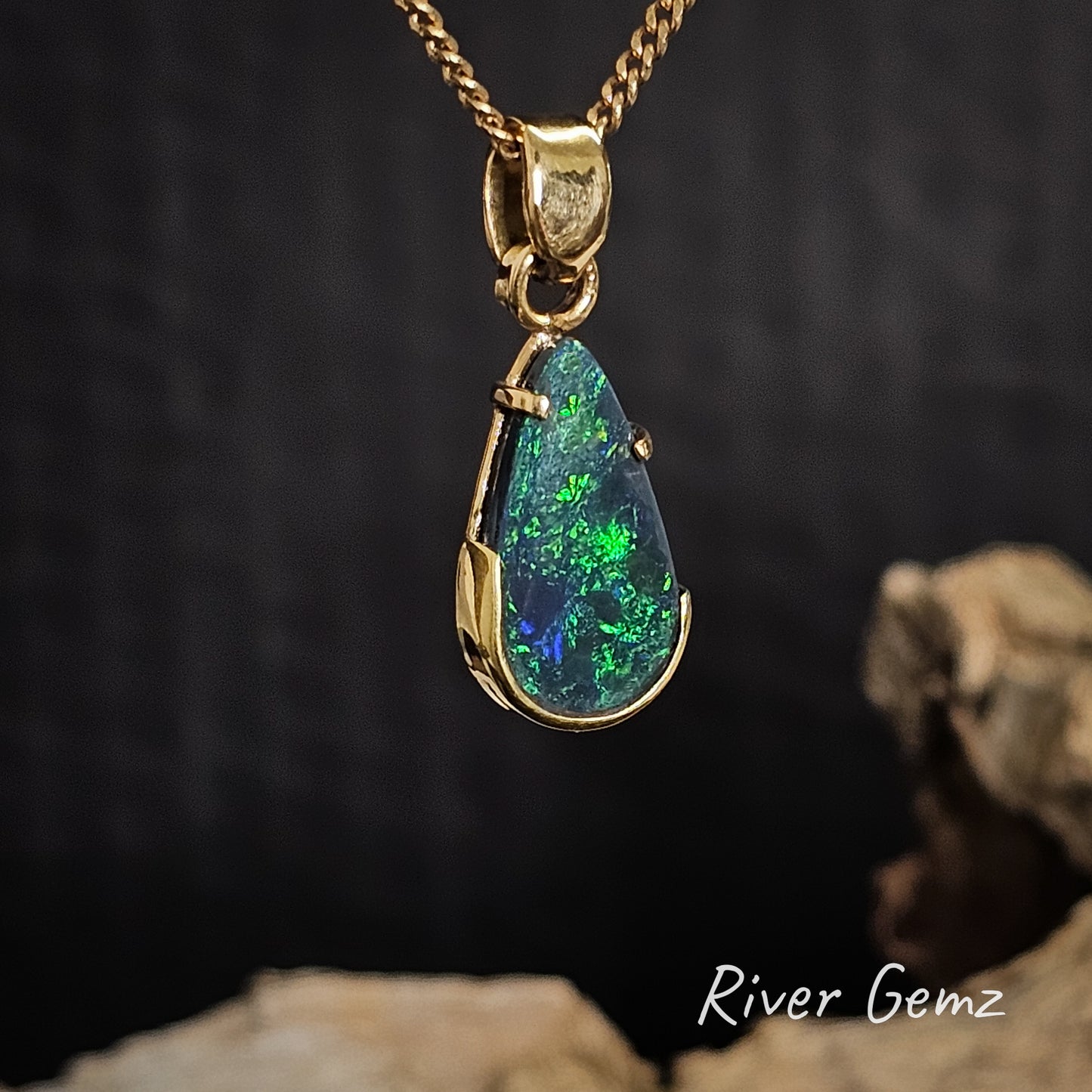 Tear drop shaped dark opal set in gold necklace on a dark background, featuring 'River Gemz' branding.