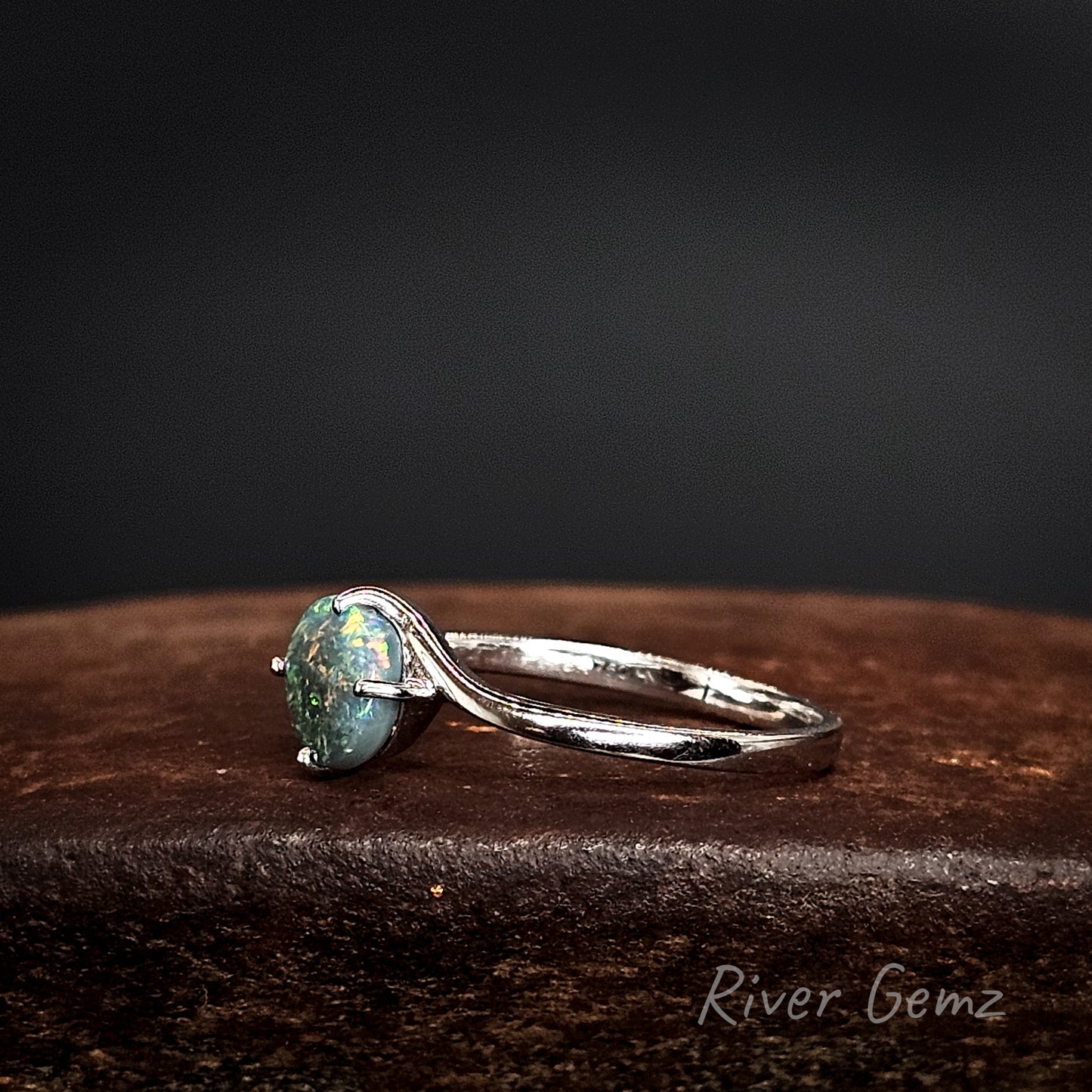 Side view shows the uniform thickness of both the ring's band and the opal on a dark surface, branded 'River Gemz'.