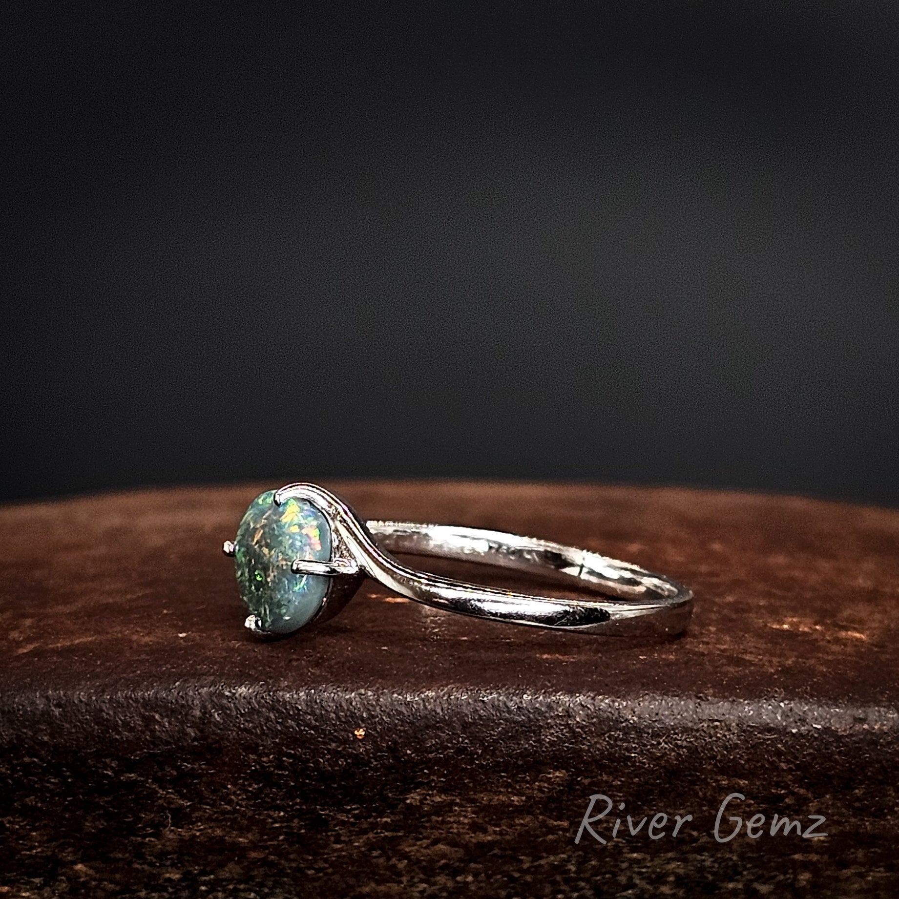 Side view shows the uniform thickness of both the ring's band and the opal on a dark surface, branded 'River Gemz'.