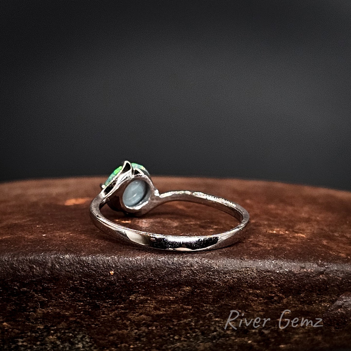 Rear view shows the dark base tone of the black opal set in the white gold ring on a dark surface, featuring 'River Gemz' branding.