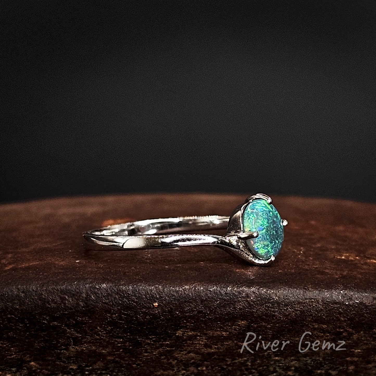 Side view of the white gold ring with claw set black opal on a dark surface, branded 'River Gemz'.