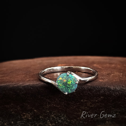 White gold ring with a round black opal stone on a dark brown surface, branded 'River Gemz'.