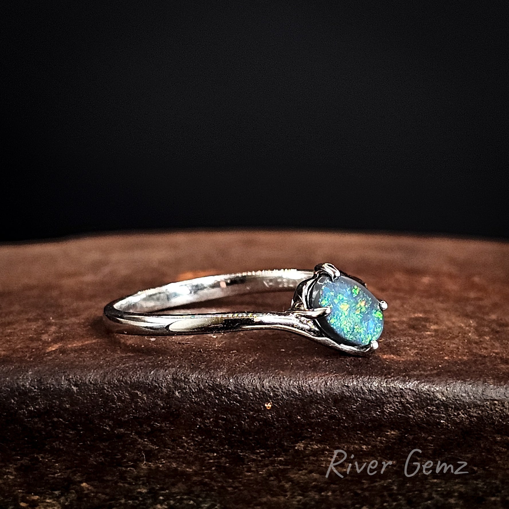 White gold ring with a claw set blue and green opal stone on a dark surface, branded 'River Gemz'.