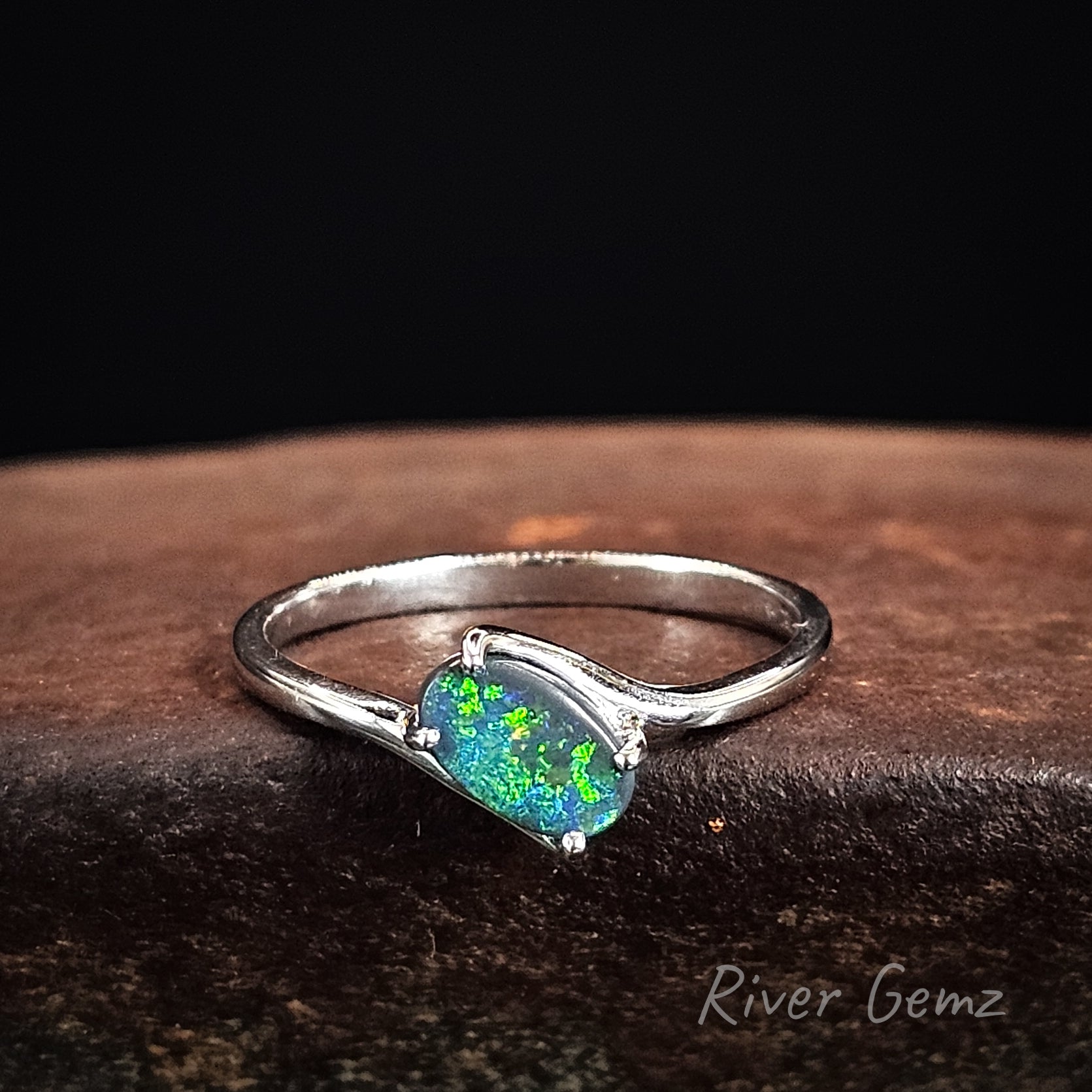 White gold ring with an oval shaped green black opal stone on a dark surface, branded 'River Gemz'.