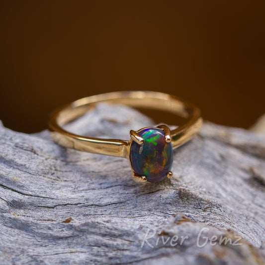 Cabochon cut, oval shaped, black opal held securely with 4 claws in the yellow gold setting. The ring is resting on a light-grey piece of driftwood.