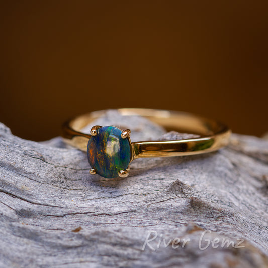 Cabochon cut, oval shaped, solid opal held securely by 4 claws in the gold ring. The ring sits on a light-grey weathered piece of wood. Opal is slightly off centre in the photo and is marginally to the left.