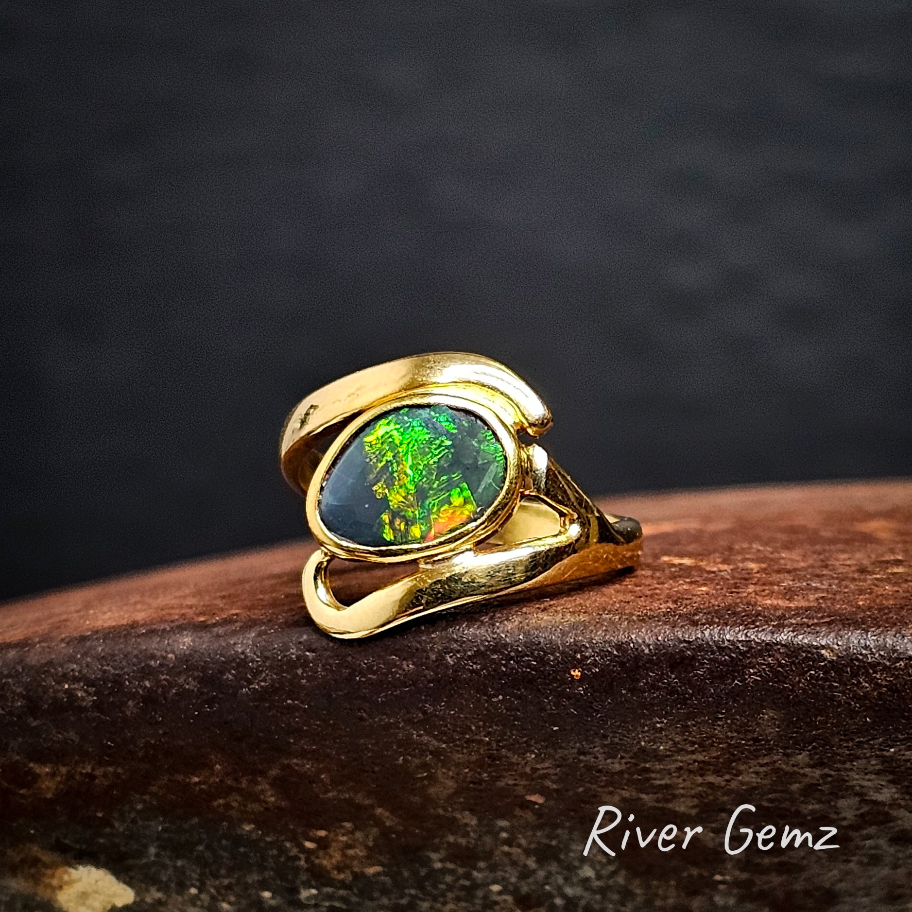 Gold ring with a green opal stone on a dark rust-like surface, branded 'River Gemz'.