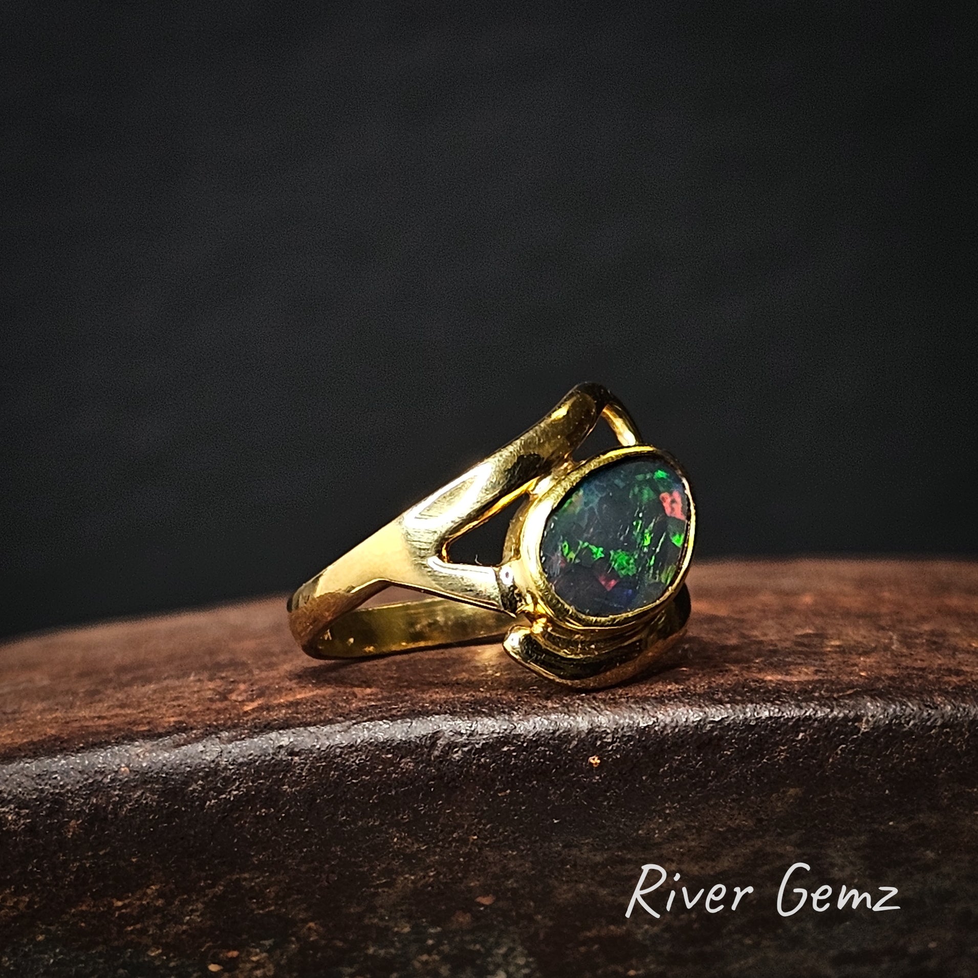 Gold ring with a black opal stone on a dark background, branded 'River Gemz'.