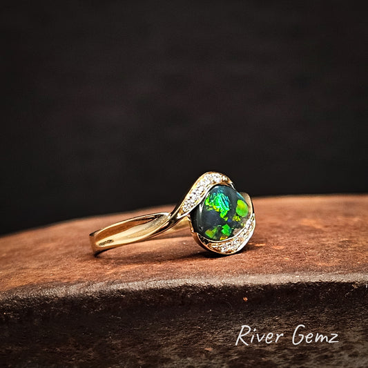 Multi-coloured black opal and diamonds in yellow gold ring on cast iron surface with 'River Gemz' branding.