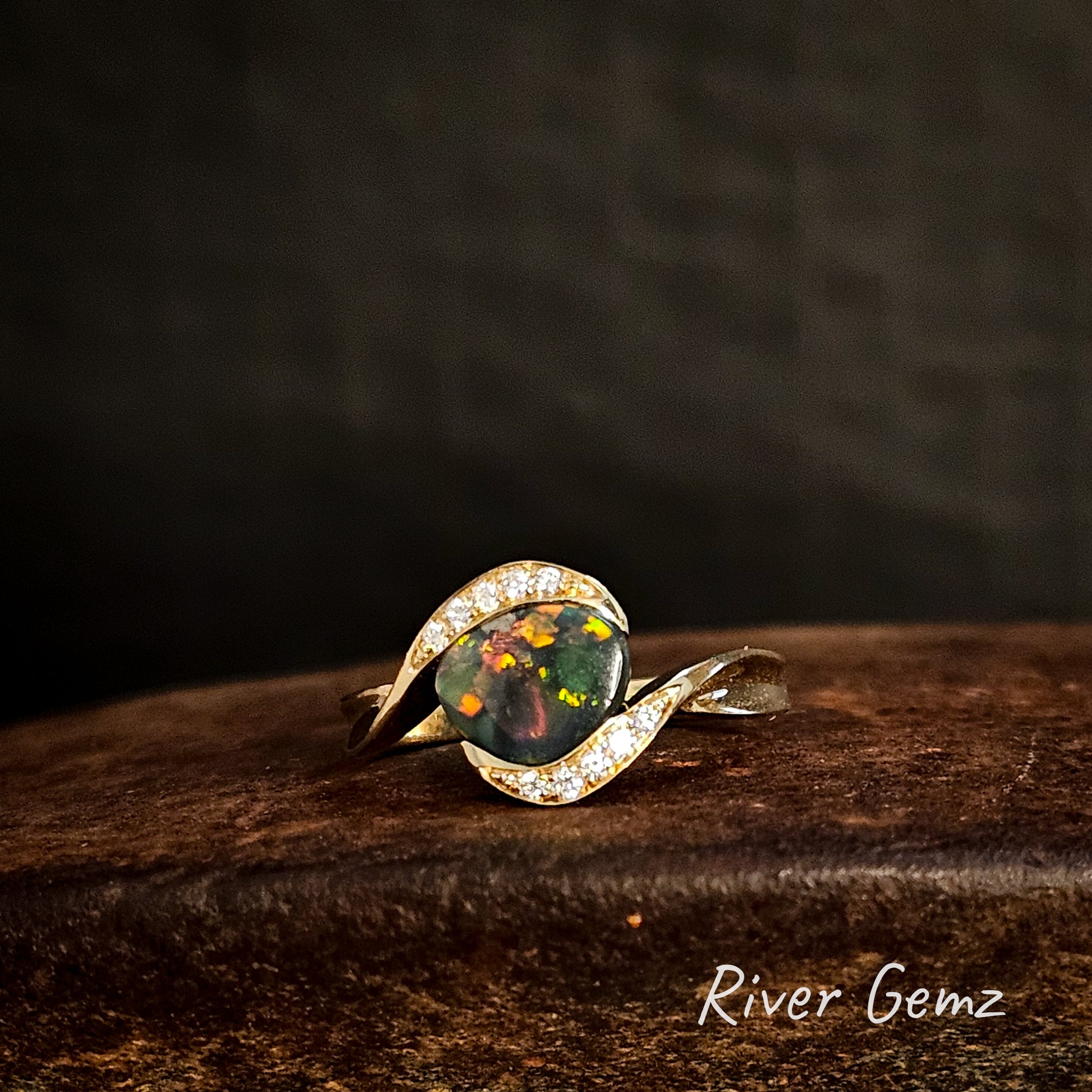 Opal ring with gold band on a dark wooden surface, 'River Gemz' brand visible