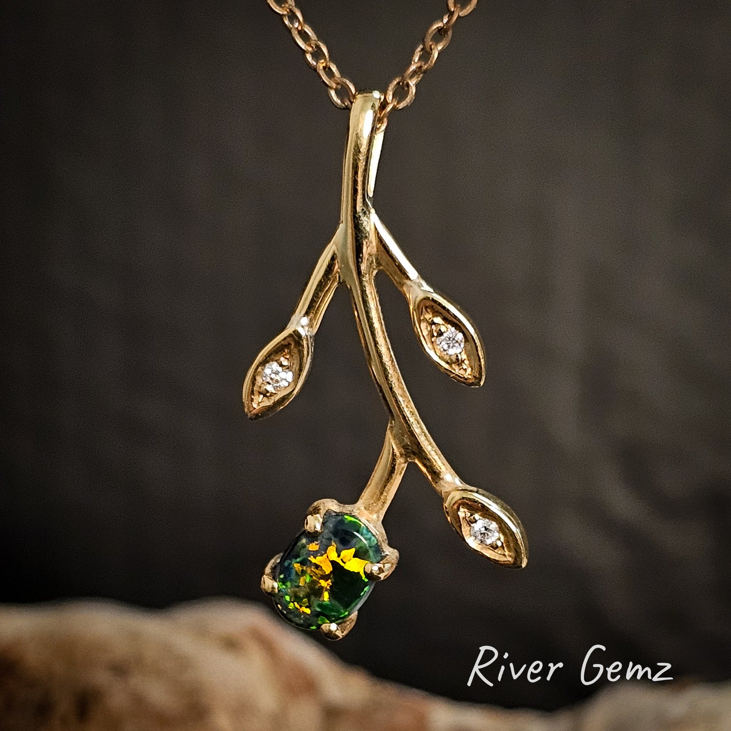 Gold branch-shaped pendant with green gemstone and diamonds on a dark background, branded 'River Gemz'.