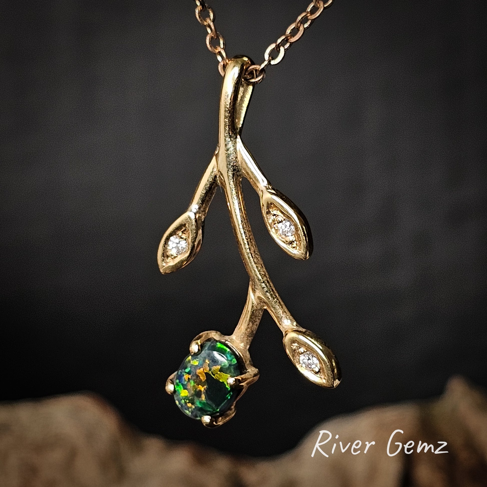 Gold branch-shaped pendant with a green gemstone on a dark background, branded 'River Gemz'.