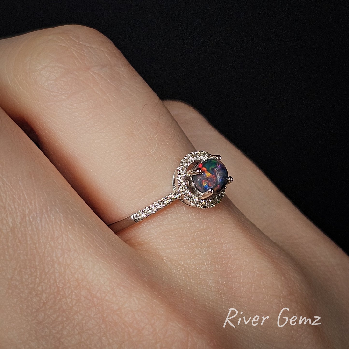 White gold ring with a colourful black opal and multiple white diamonds on a hand, branded 'River Gemz'.