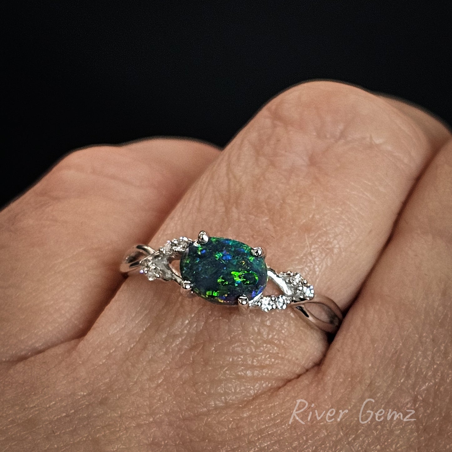 Gold ring with a green dark opal and diamonds on a finger, branded 'River Gemz'.