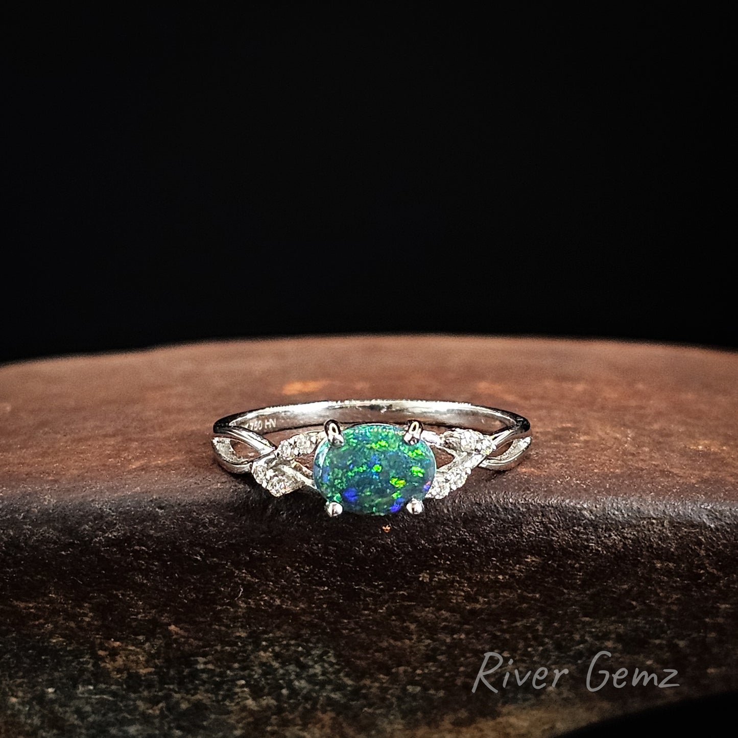 White gold ring with an oval shaped black opal and diamonds on a dark surface, branded 'River Gemz'.