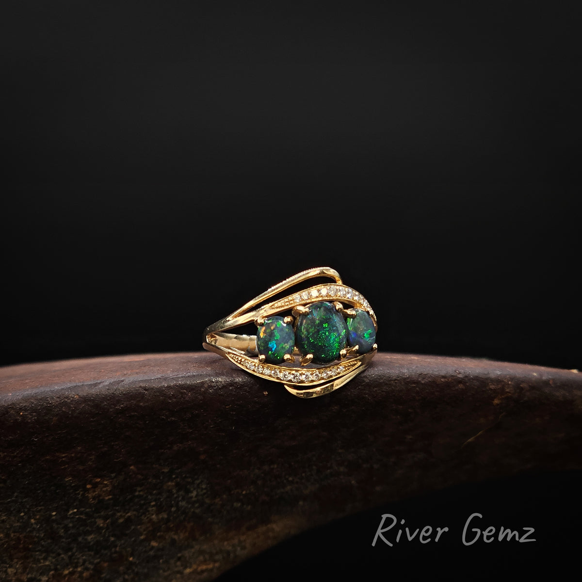 Three dark green oval shaped opals and 2 lines of channel set white diamonds set in a gold ring on a brown surface against a dark background.
