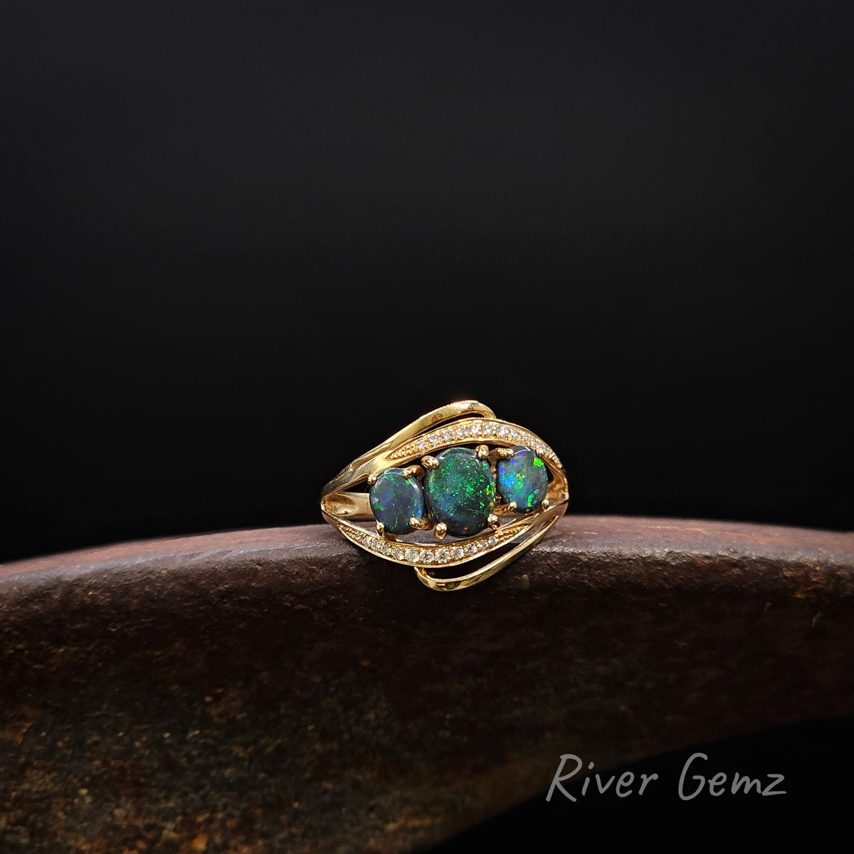 Three dark green oval shaped opals and 2 lines of channel set white diamonds set in a gold ring on a brown surface against a dark background.