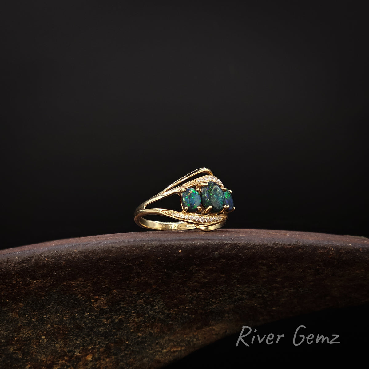 Three dark green oval shaped opals and 2 lines of channel set white diamonds set in a gold ring on a brown surface against a dark background.
