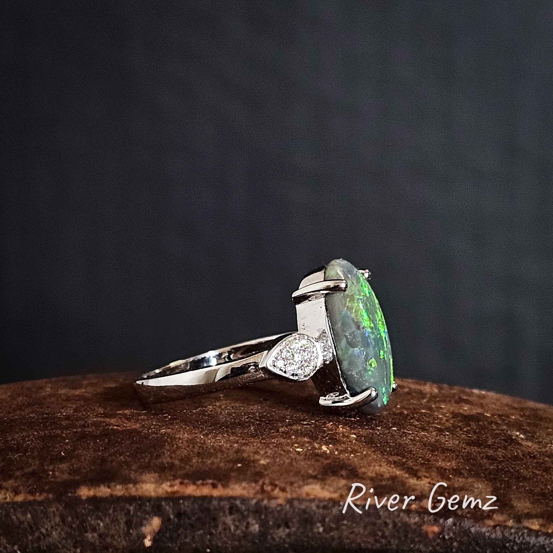 Side view of the ring showing the thickness of the oval shaped solid black opal on a dark background, branded 'River Gemz'.