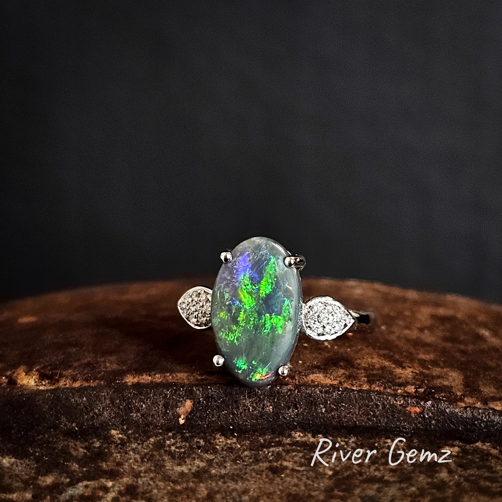 Ring with an oval shaped green black opal and pave diamonds on the shoulders on a dark background, featuring the brand 'River Gemz'.