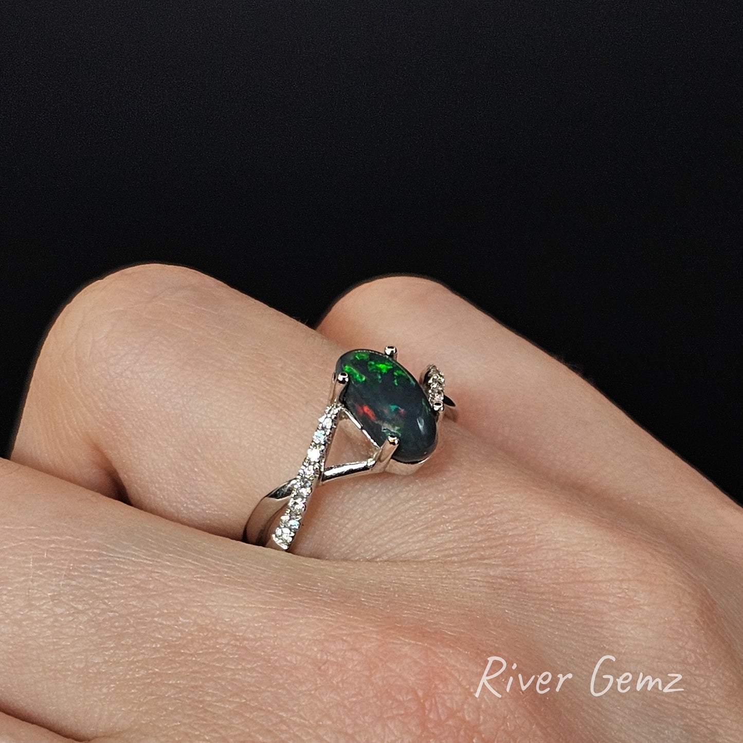 Ring with a green black opal and diamonds on the shoulders on a hand, brand 'River Gemz' visible.