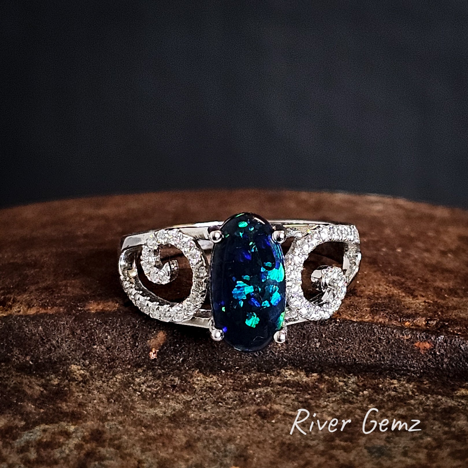 White gold ring with a dark blue and green black opal on a iron shoe anvil, branded 'River Gemz'.