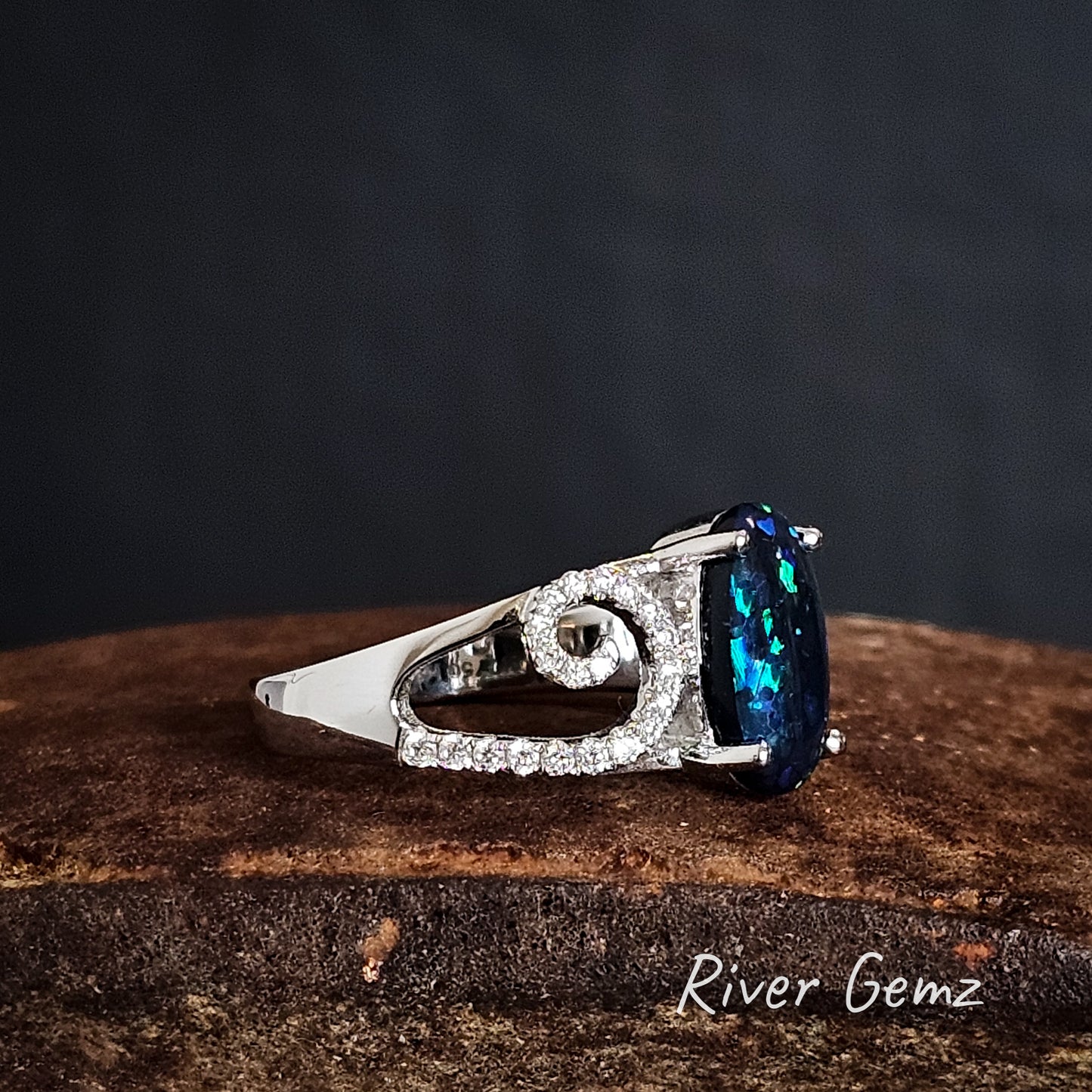 Side view of the white gold ring with a dark blue oval shaped opal and a swirl of diamonds on the rings shoulder on a rust coloured surface, branded 'River Gemz'.