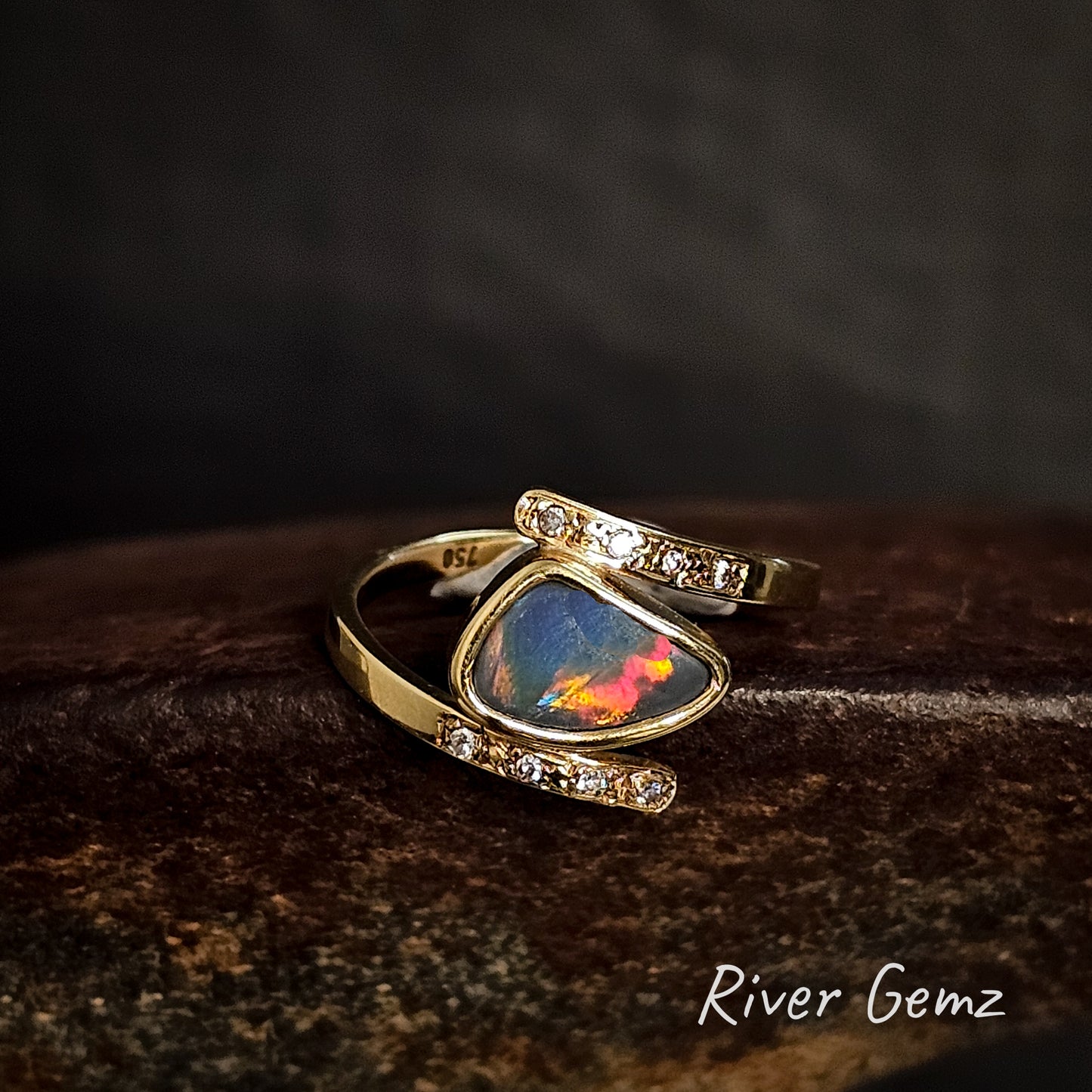 Gold ring with a dark opal stone on a dark brown surface, featuring 'River Gemz' branding.
