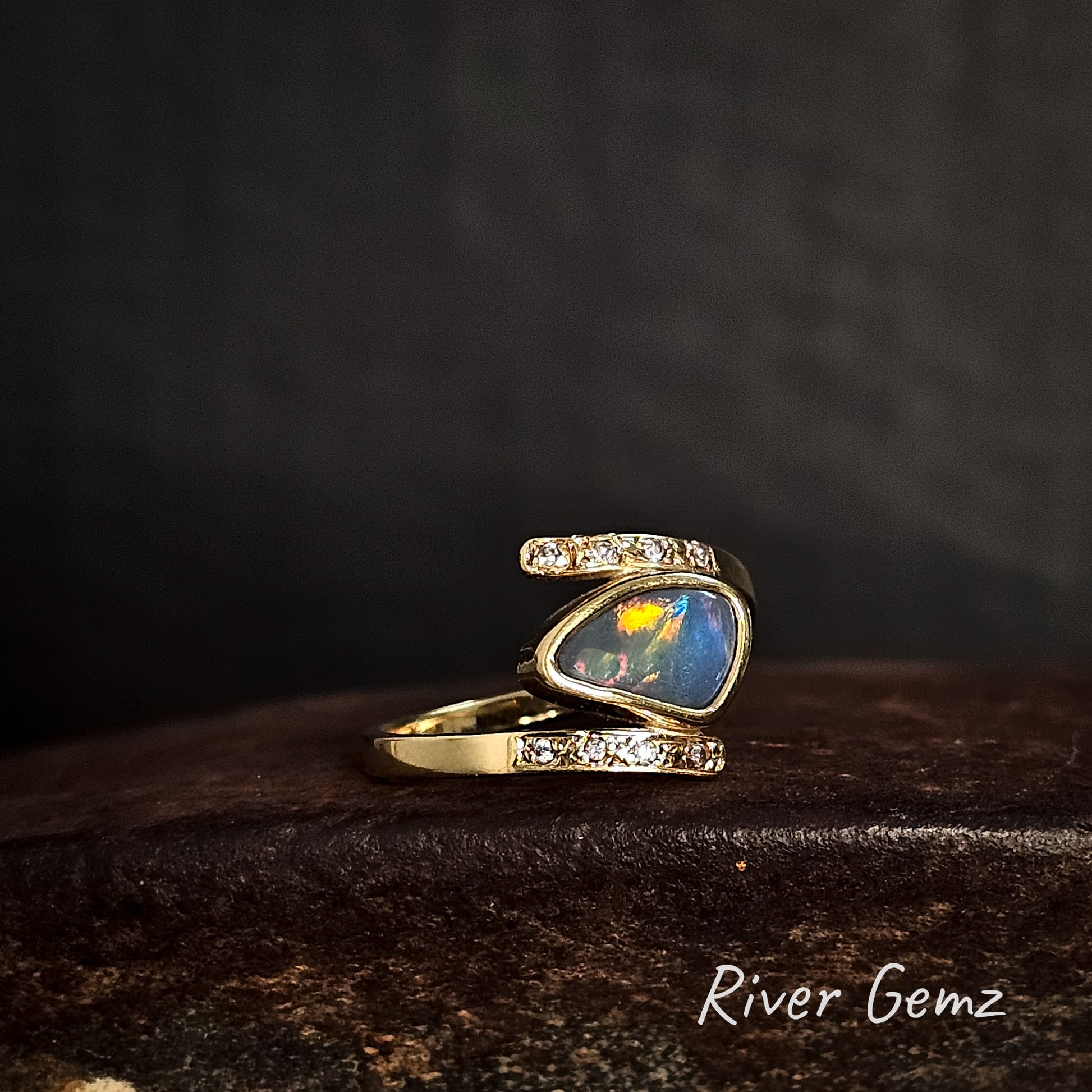 Gold ring with opal stone and diamonds on a dark surface, 'River Gemz' brand visible.
