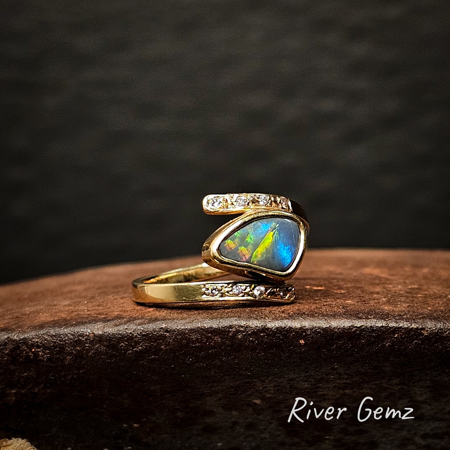 Gold ring with diamonds and a free form cut colourful opal stone on a dark surface, branded 'River Gemz'.