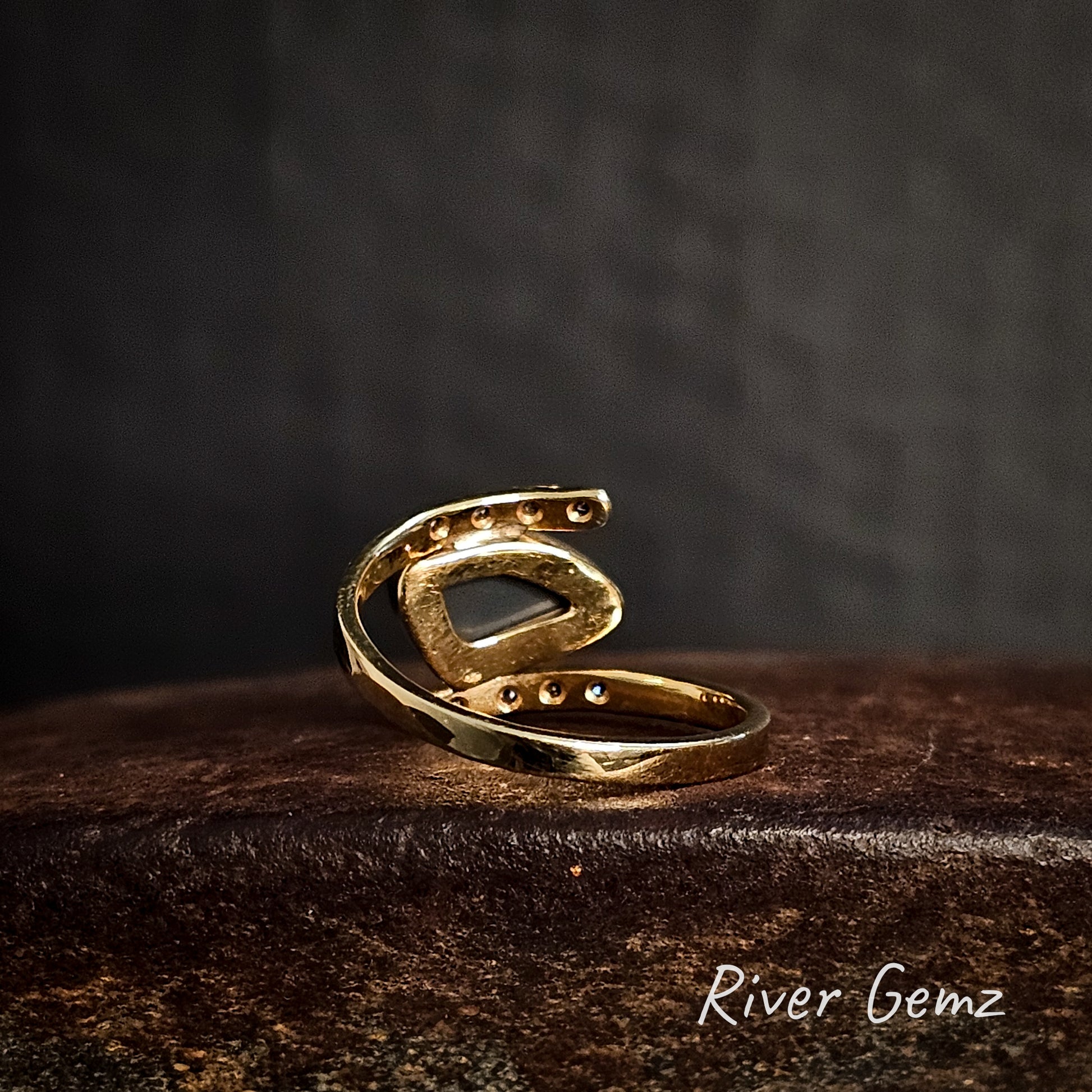 Rear view of the gold ring showing the dark tone of the black opal set in it on a dark surface with 'River Gemz' branding.