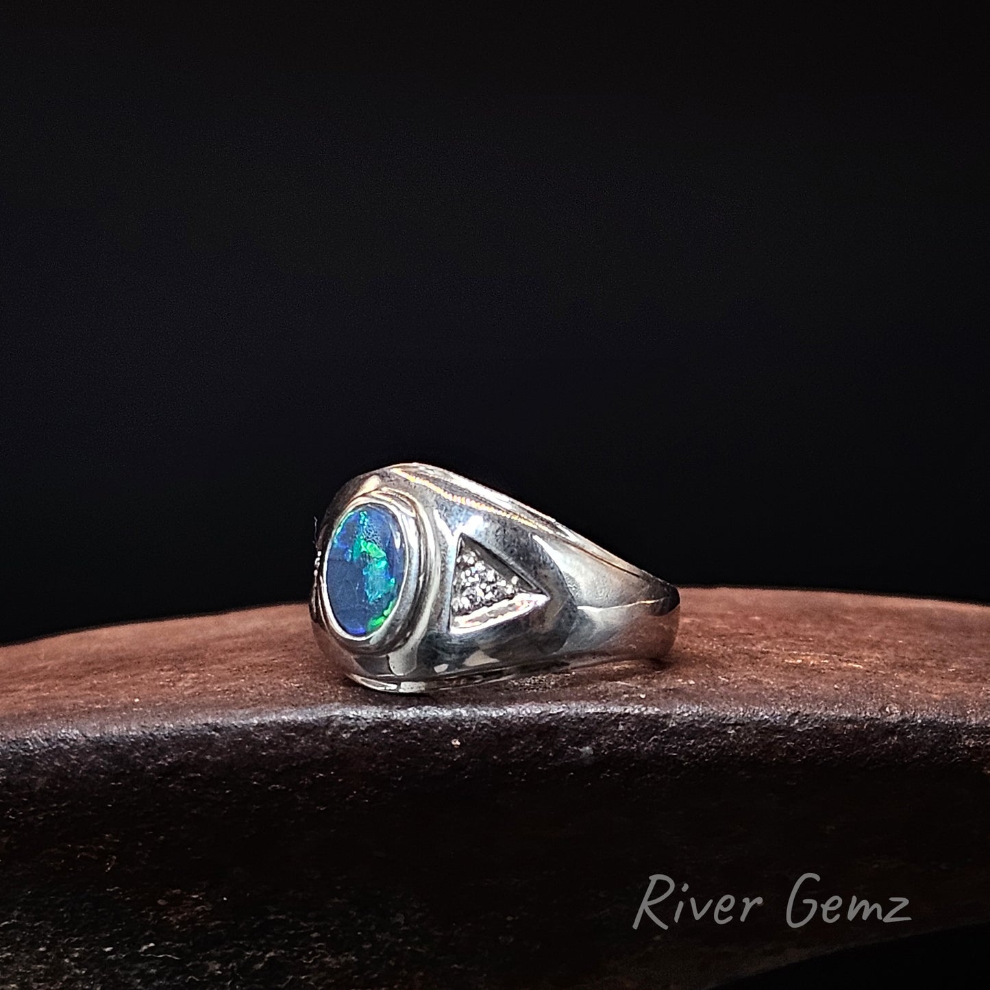 A sterling silver signet ring featuring a round iridescent blue-green opal gemstone and 3 small diamond accent stones, resting on a dark wooden surface against a black background.