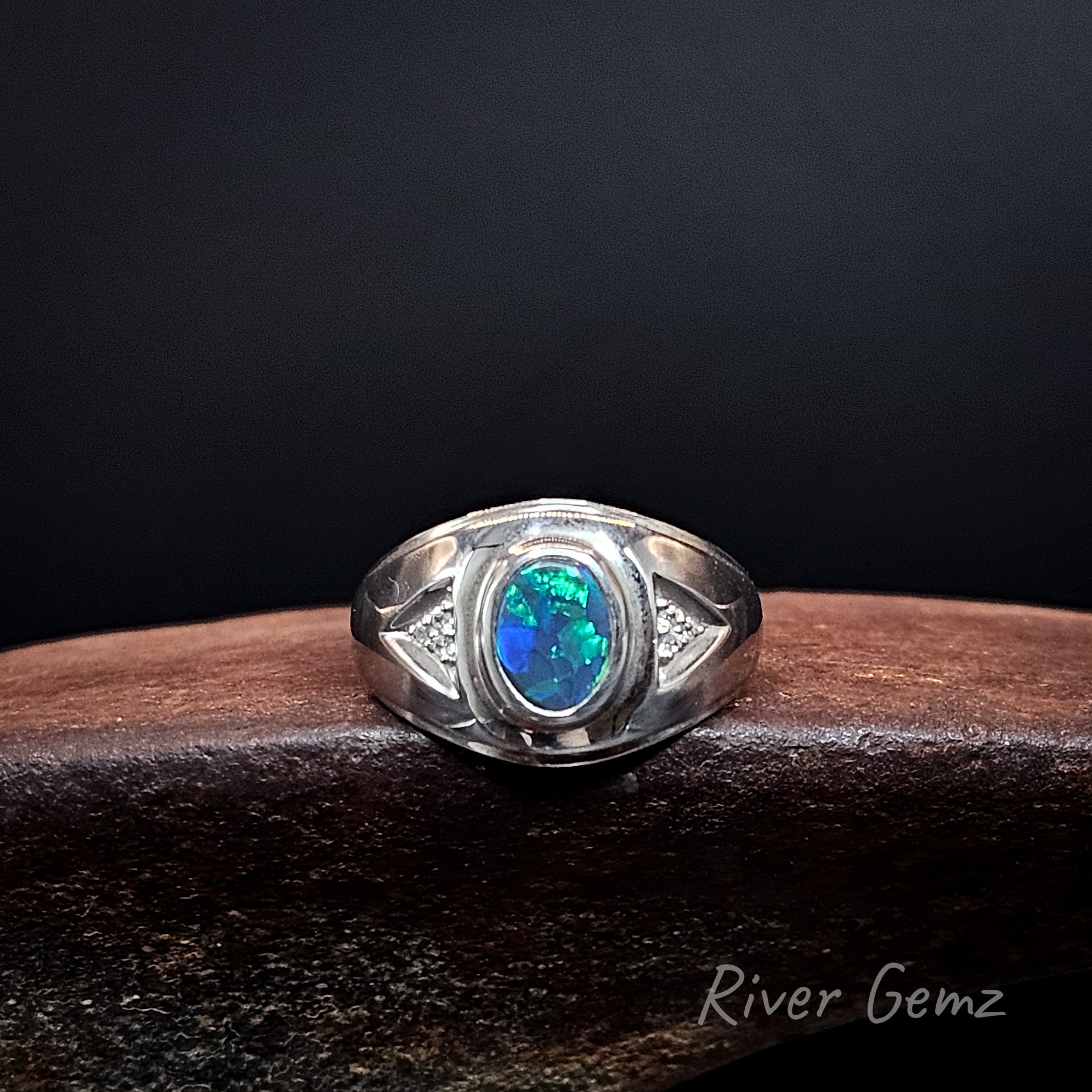 A sterling silver ring featuring an oval-cut blue opal gemstone, displayed on a dark wooden block against a black background. The brand name 'River Gemz' is visible as a watermark in the bottom right corner.