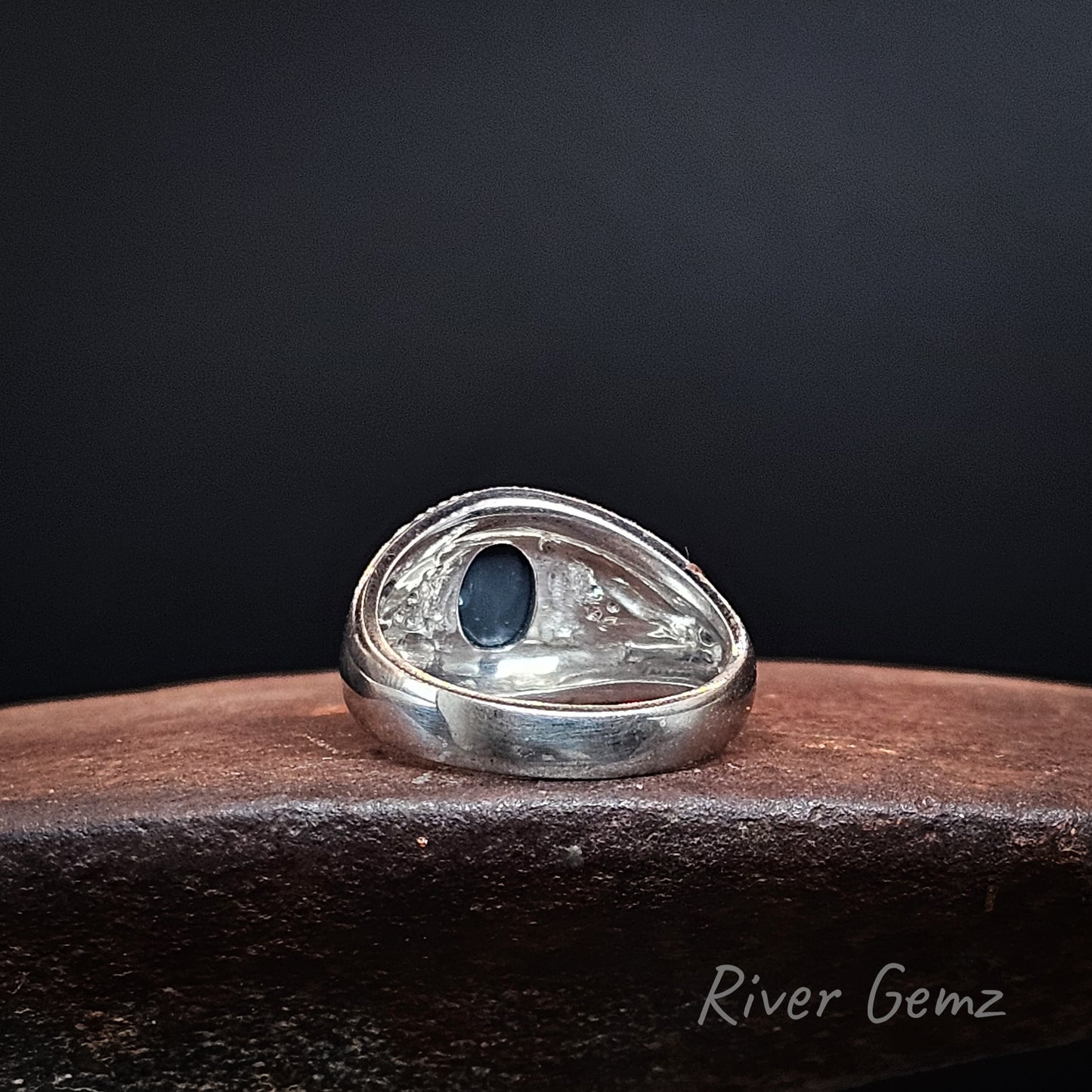 Rear view of silver-toned ring showing the dark base tone of the included oval shaped black opal, displayed on a dark brown wooden surface against a black background. The brand name 'River Gemz' is visible in the bottom right corner.