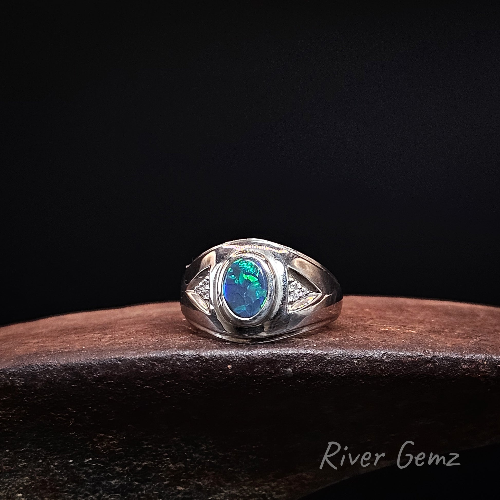 A sterling silver ring featuring an oval-cut blue opal gemstone, displayed on a dark wooden surface against a black background. The brand name 'River Gemz' is visible in the bottom right corner.