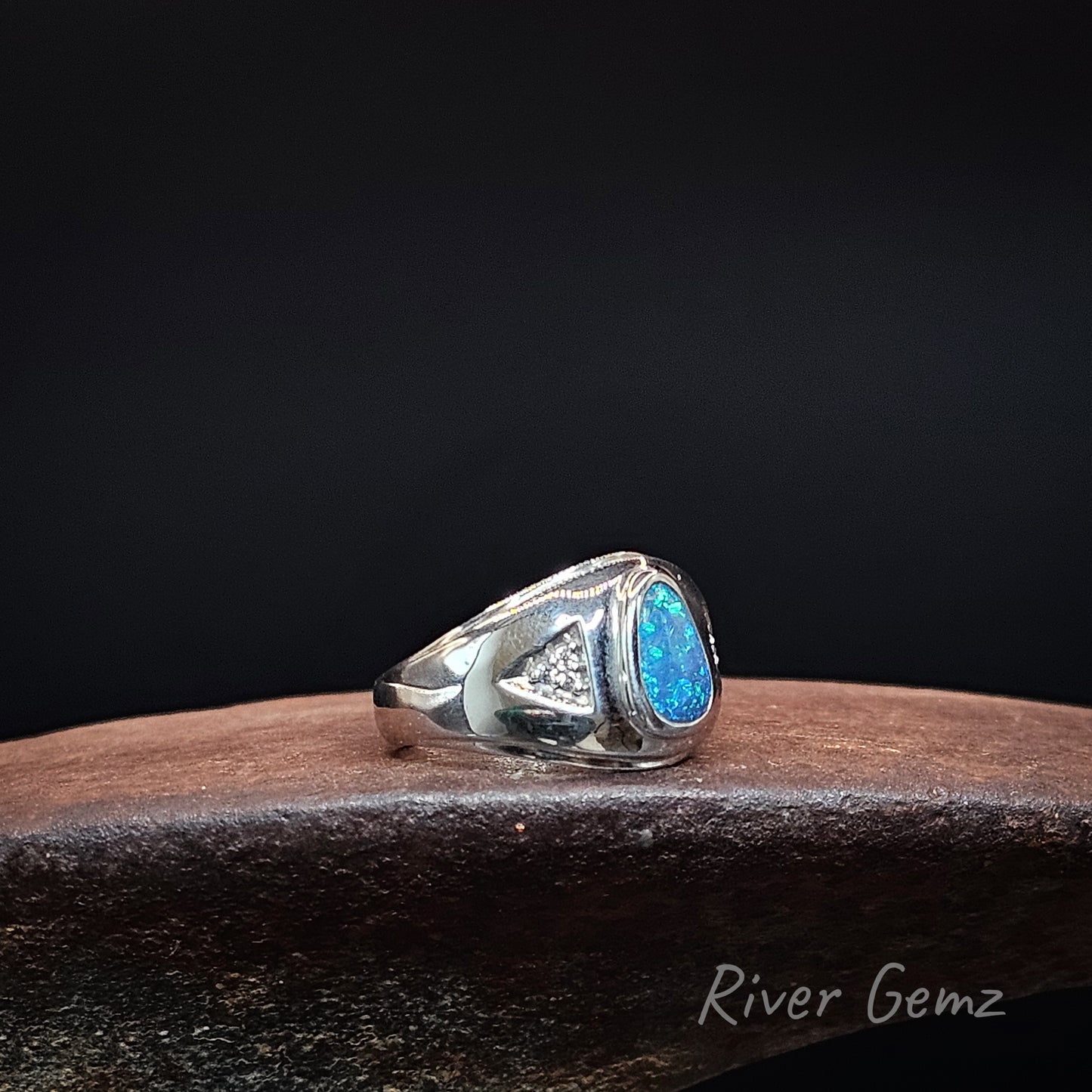 A sterling silver men's signet ring with a vibrant blue and green opal gemstone and a small triangle of diamonds, resting on a dark wooden surface against a black background.
